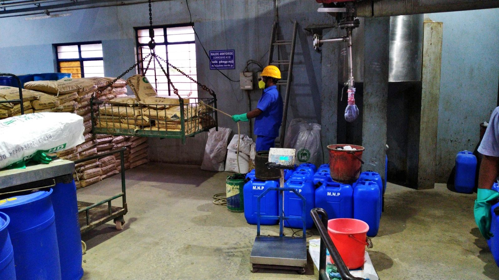 Worker handling a suspended material cage near stacked bags and blue containers inside the factory.