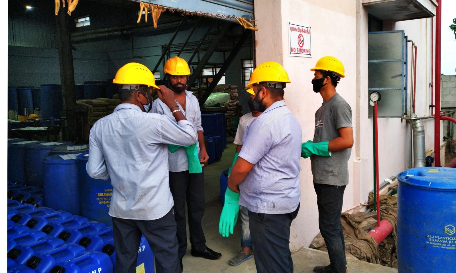 Staff wearing protective equipment beside stored blue containers at the factory entrance.